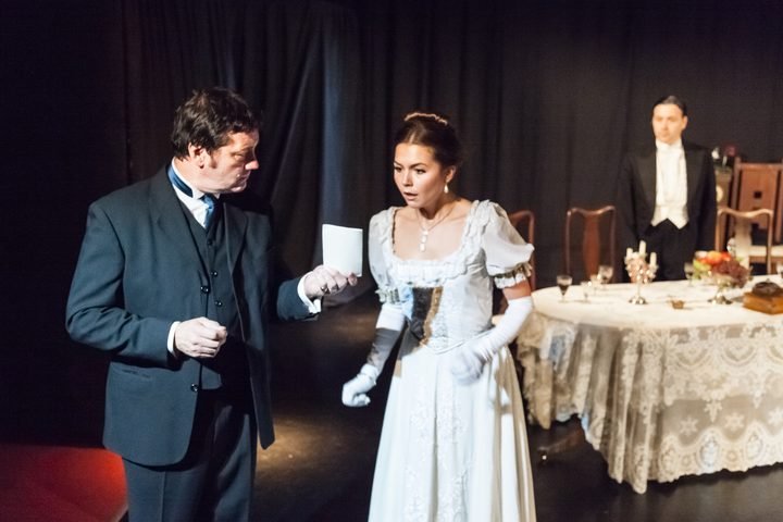 A scene from a 2012 OVO Theatre production of An Inspector Calls: the Inspector holds out a photograph to Sheila while Gerald watches.