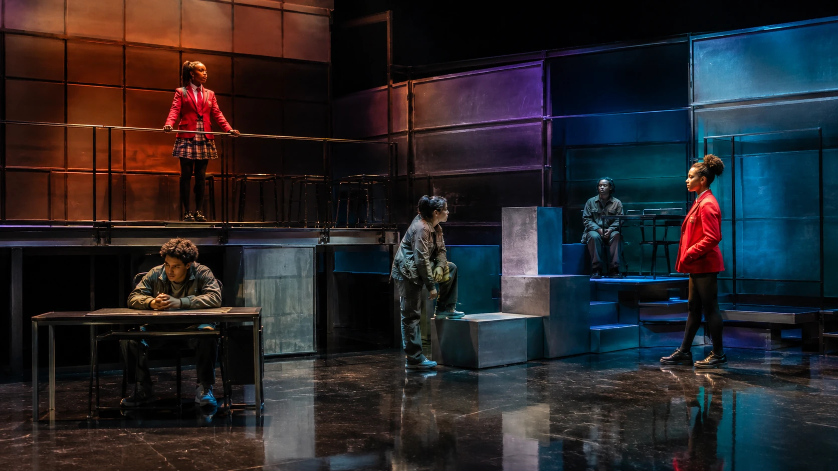 A flexible live-theatre set divided into levels, desks, blocks and open acting areas under red, teal and purple light.